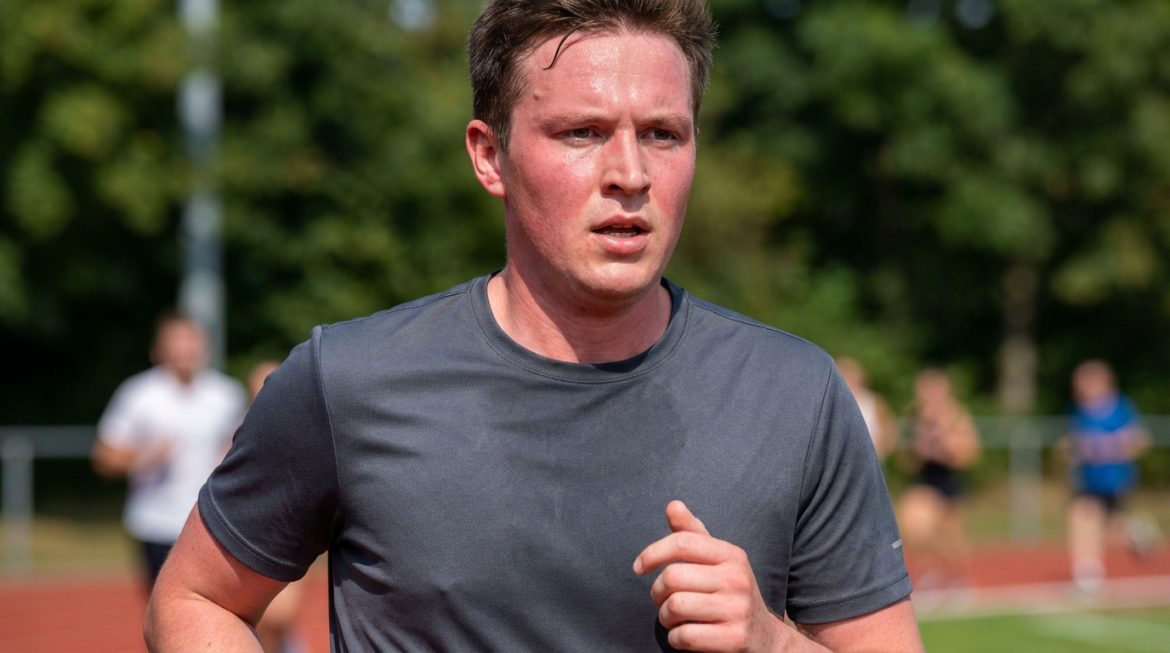 Un jeune homme court sur une piste athlétique, son visage est très rouge et couvert de sueur sous le soleil.