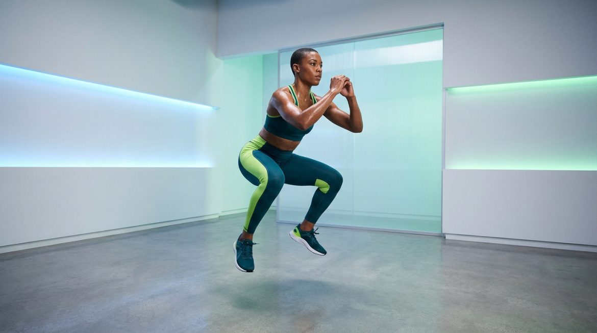 Femme en tenue de sport vert émeraude et vert lime réalisant un squat jump dynamique. Transpiration visible, fond moderne et lumineux.